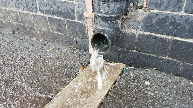 Downspout outlet emptying rainwater onto a board at the base of a wall